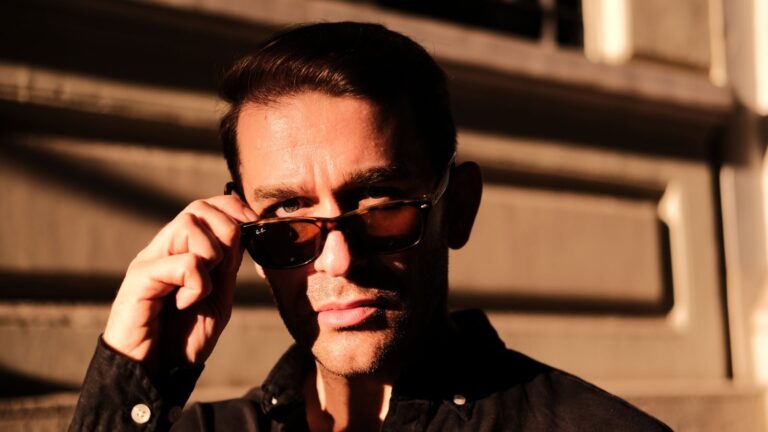 A stylish man adjusts his sunglasses under sunset lighting, casting dramatic shadows on an urban setting.
