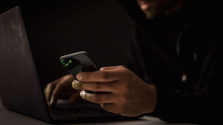 Crop anonymous ethnic male cyber spy with cellphone and netbook hacking system in evening