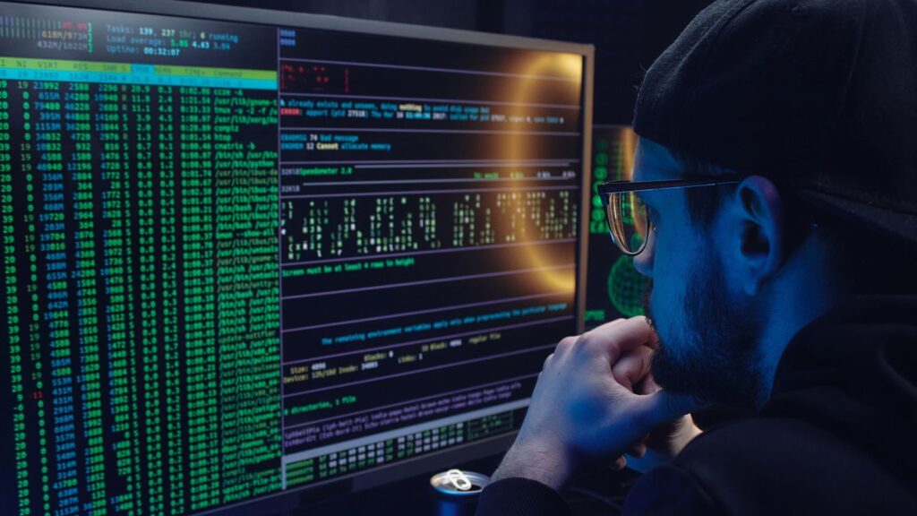 Man intently working on computer programming with code displayed on dual monitors in a dimly lit room.