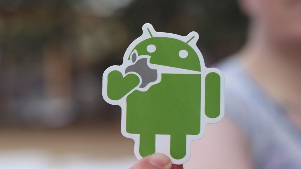 Close-up of an Android sticker, humorously holding an apple logo, with a blurred outdoor background.