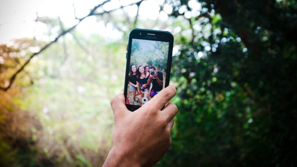 A hand holds a smartphone capturing a selfie of friends in a lush outdoor setting.