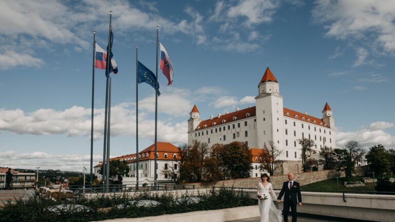 A newlywed couple holding hands in front of Bratislava Castle, Slovakia.