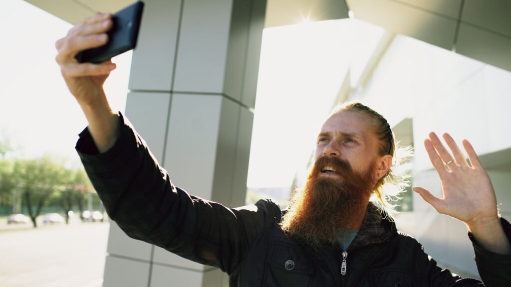 Bearded man taking a selfie outside modern architecture on a sunny day.