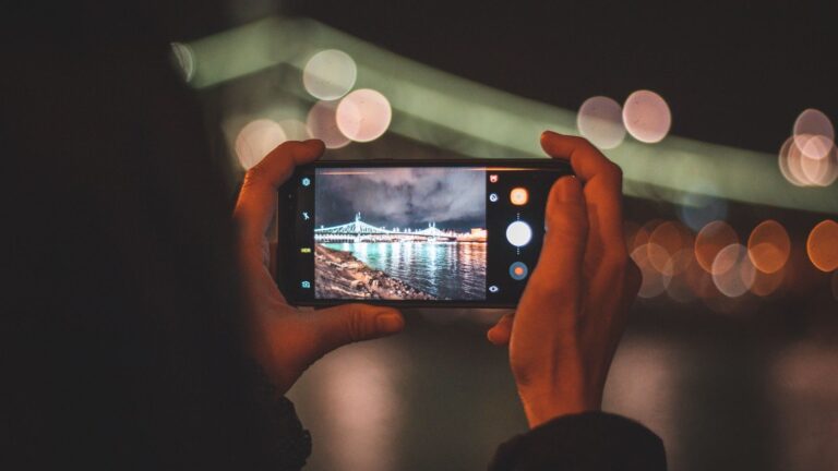 Hands holding a smartphone capturing a cityscape at night with colorful bokeh lights.