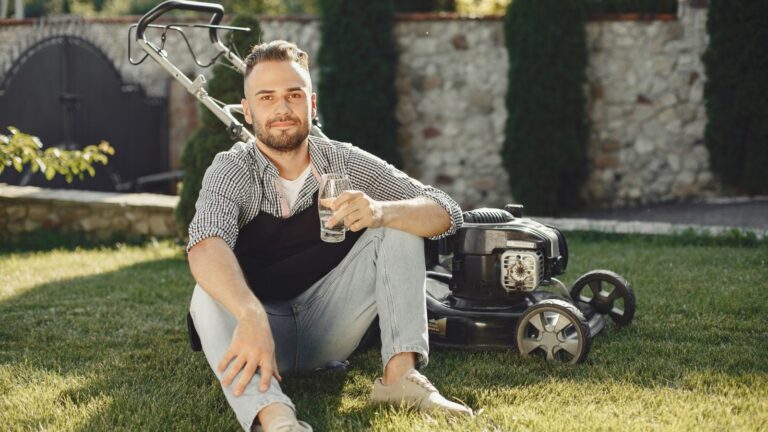Man sitting on grass with drink, beside lawn mower on a sunny day.