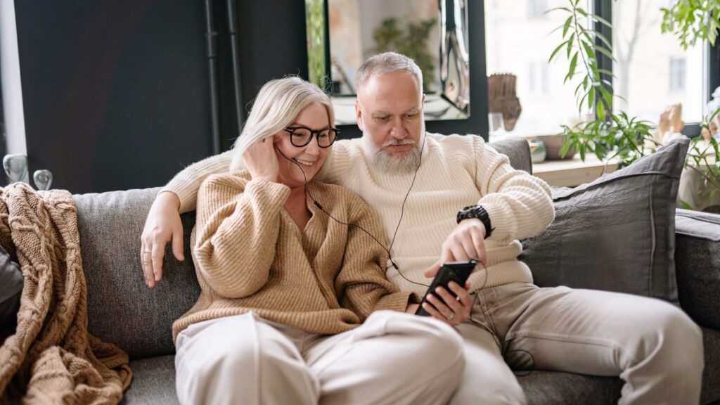 Senior couple enjoying music on a cozy couch, sharing headphones in a warm home setting.