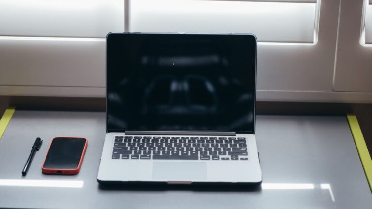 A minimalist desk setup featuring a laptop, smartphone, and pen under soft window light.