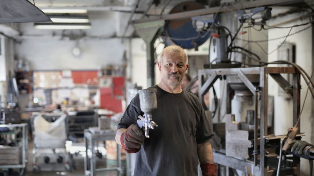 Pleased senior white hair craftsman in heavy duty gloves showing sandblaster and looking at camera