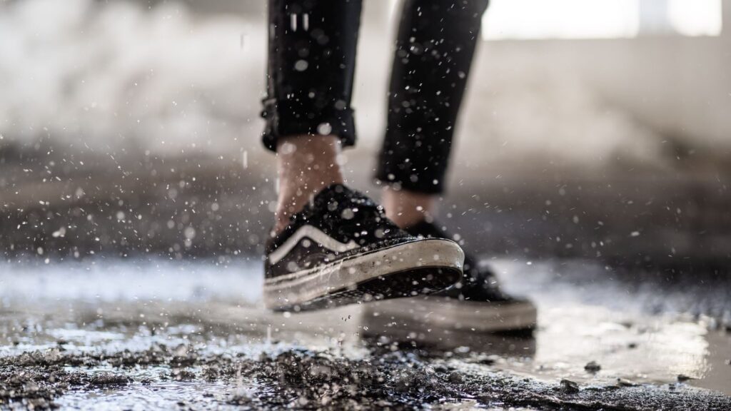 Crop anonymous person in rag sneakers strolling on wet ground while stomping through puddles and creates splashes on rainy day