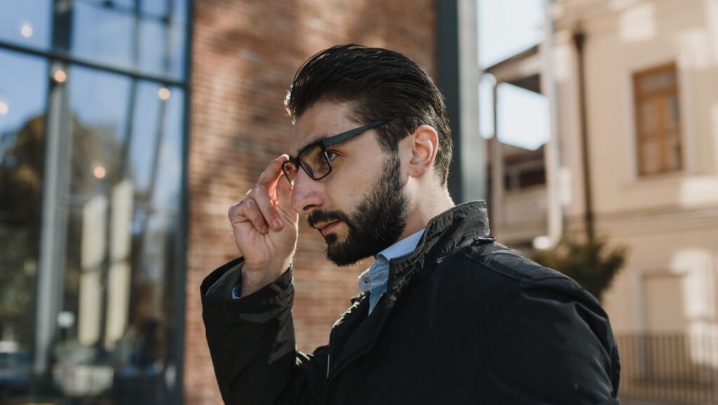 Profile of a bearded businessman adjusting his glasses outside a modern building on a sunny day.