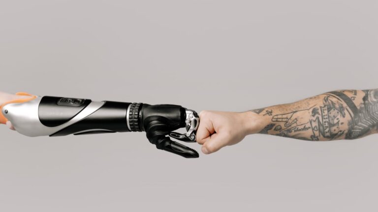 Close-up of a tattooed arm fist bumping with a robotic prosthetic arm in studio setting.