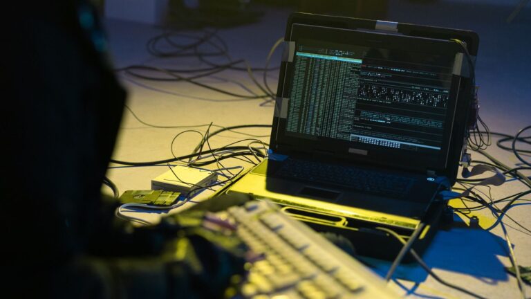 A person typing on a keyboard connected to a laptop showing code, surrounded by wires.