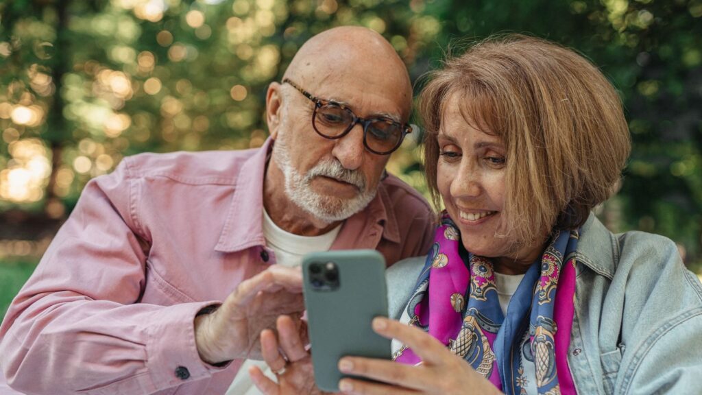 Happy senior couple using smartphone in a park, enjoying sunny outdoors.