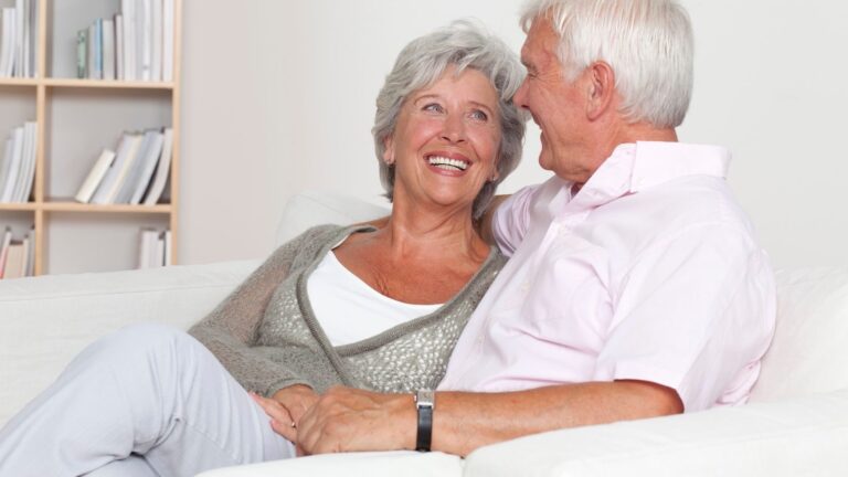 Smiling senior Caucasian couple sitting comfortably on a sofa in a bright room full of books.