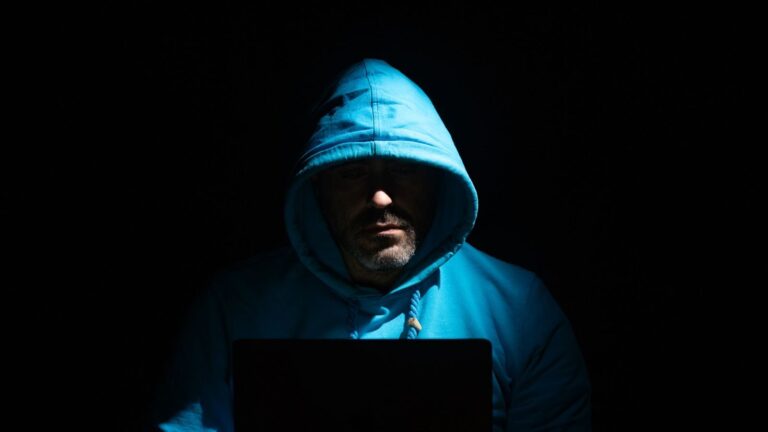 A man in a blue hoodie focused on a laptop in a dark room, conveying secrecy and intrigue.