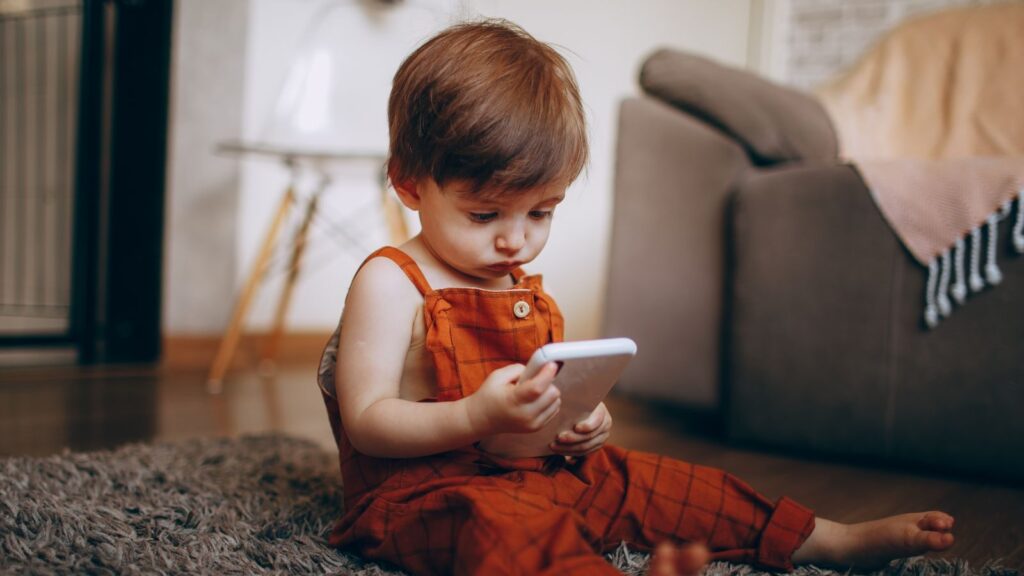 A cute toddler in orange overalls playing with a smartphone on a cozy carpet indoors.