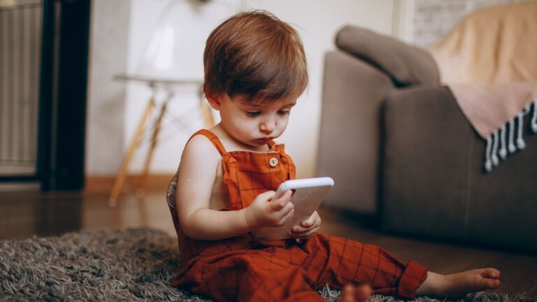 A cute toddler in orange overalls playing with a smartphone on a cozy carpet indoors.